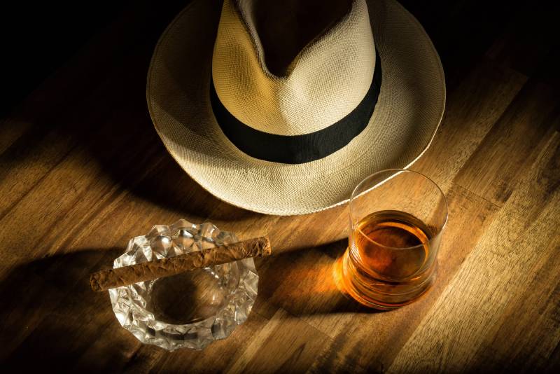 Cigare artisanal sur cendrier en cristal avec whisky âgé et chapeau Dominicain en fond.